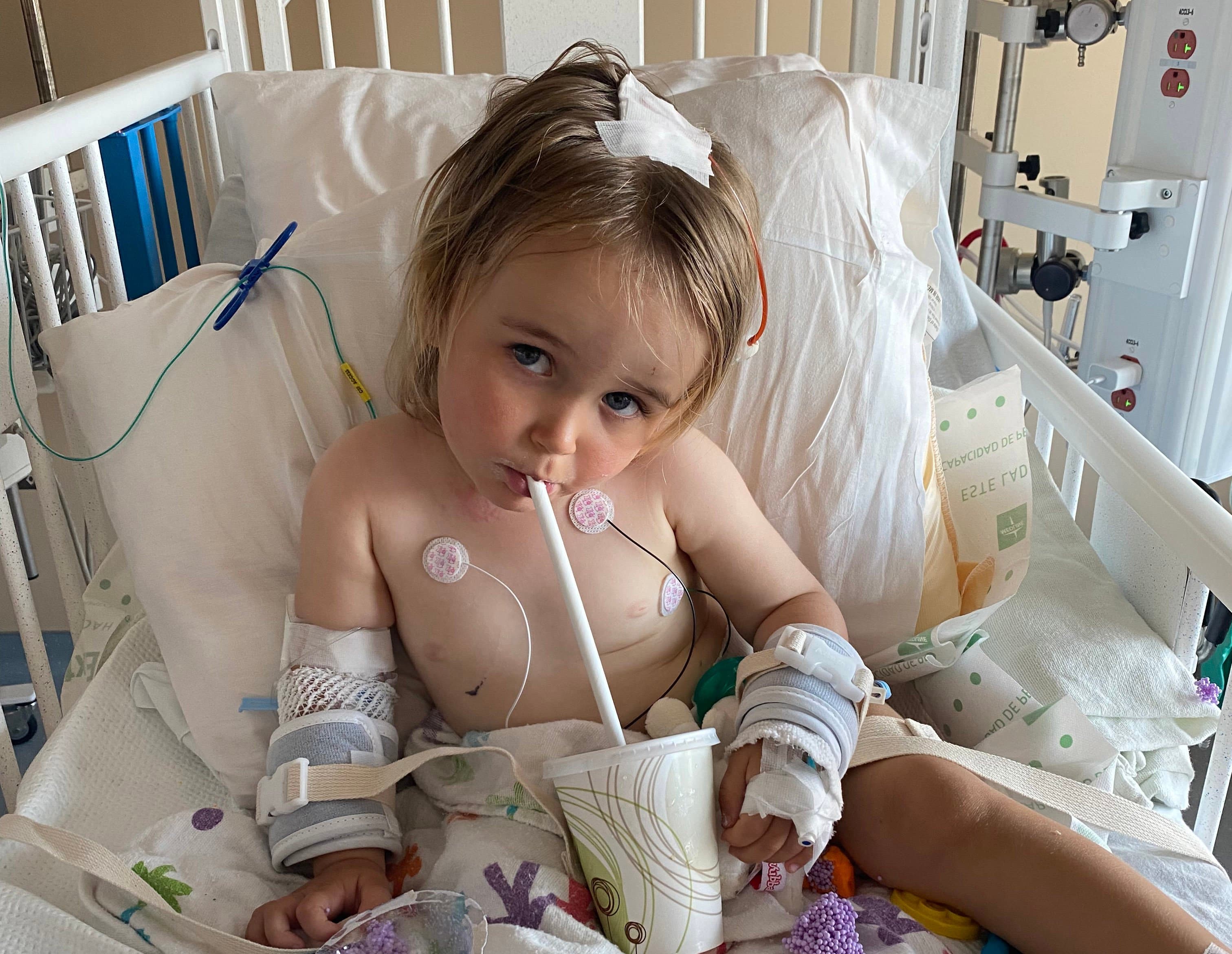A toddler in a hospital bed with a chest monitor and head bandage sipping from a sippy cup