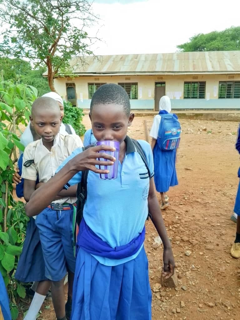 Schoolgirl in a blue uniform drinking clean water provided through an Outreach Program Waterpoint, with classmates gathered around.